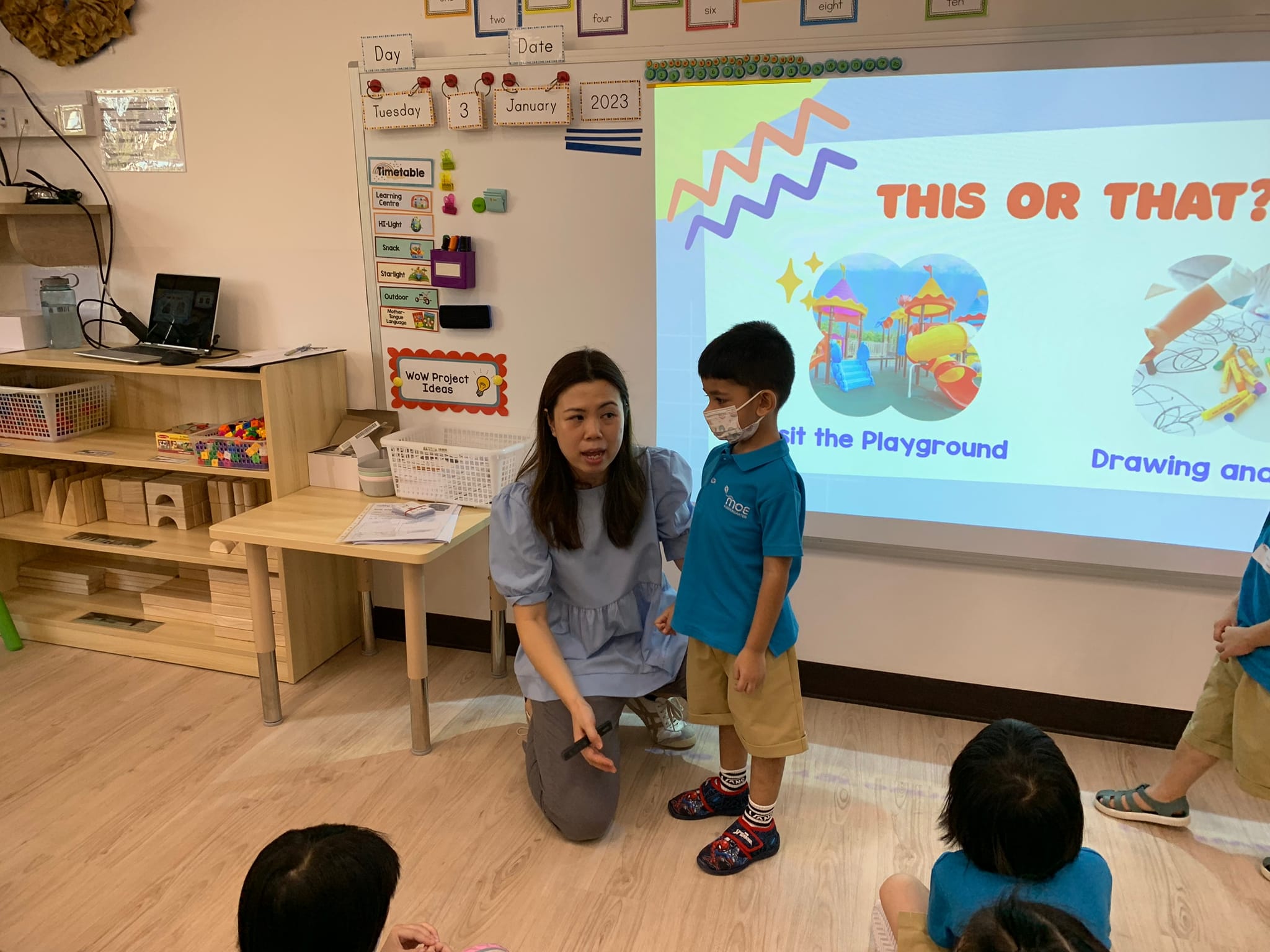 A teacher kneels beside a masked young boy in a colorful classroom with a "This or That?" slideshow projected behind them.