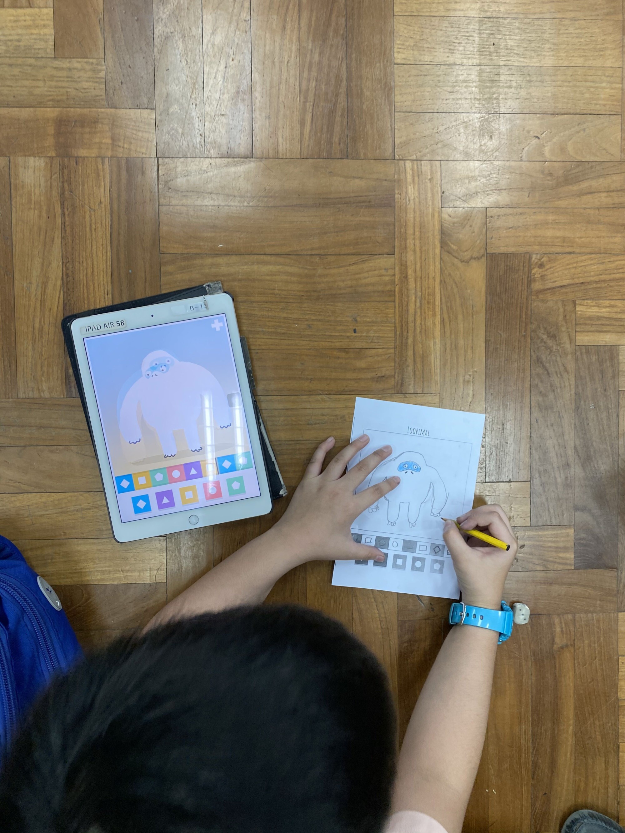 Child drawing a yeti character on paper using a tablet showing the same yeti as a reference, on a wooden floor.