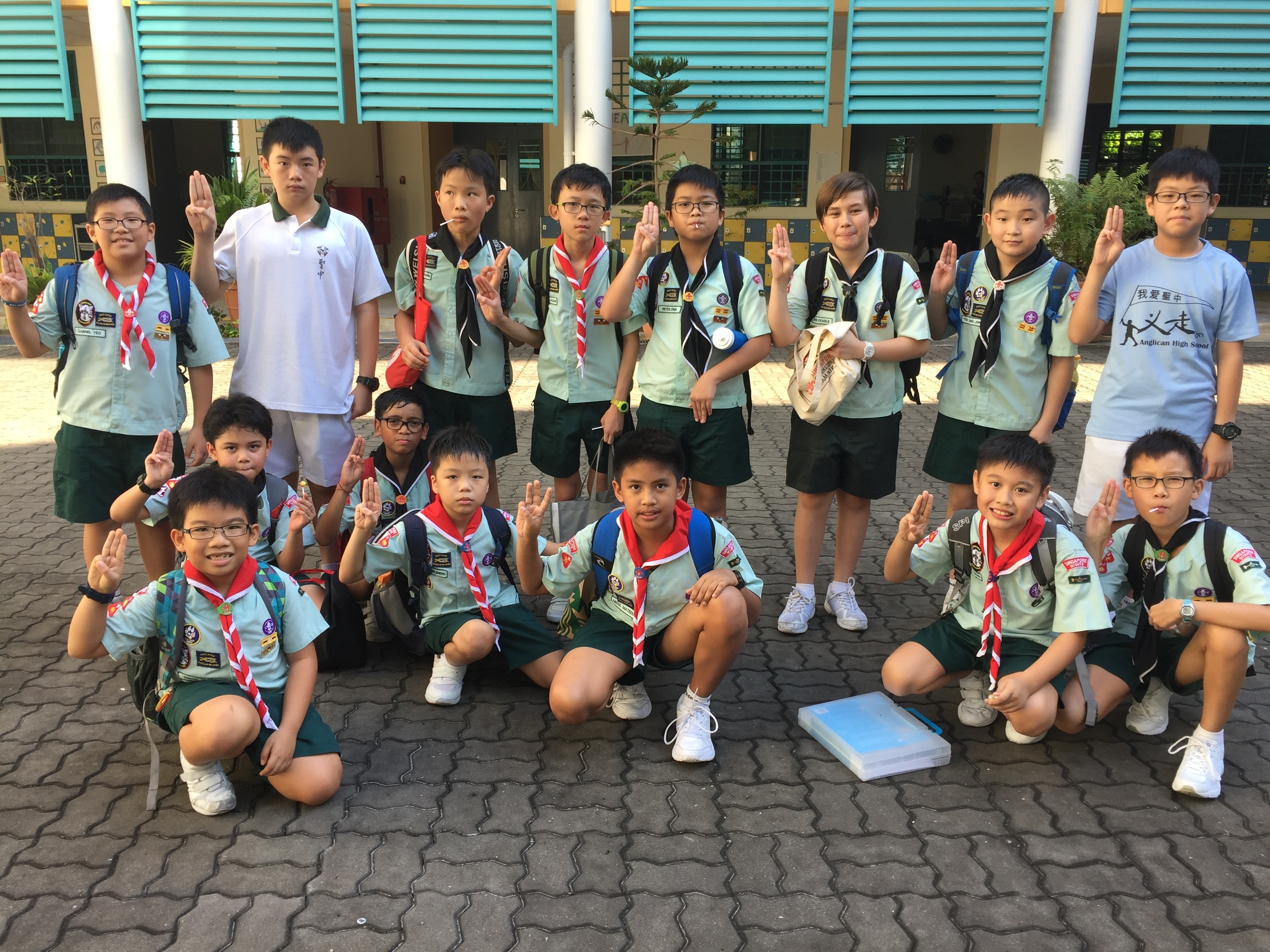 Group of young scouts in light green uniforms and red neckerchiefs posing with three-finger salute outside a school building.
