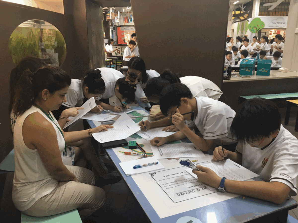 Students in white uniforms lean over a table writing on papers, guided by an adult woman in a busy indoor setting.