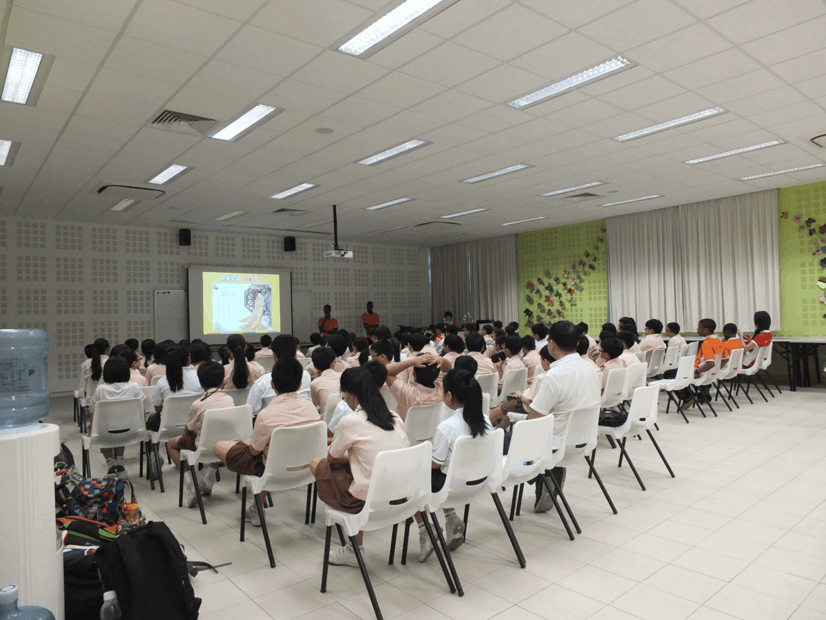 Students in pink uniforms sit on white chairs in a hall, facing a projected presentation screen at the front.