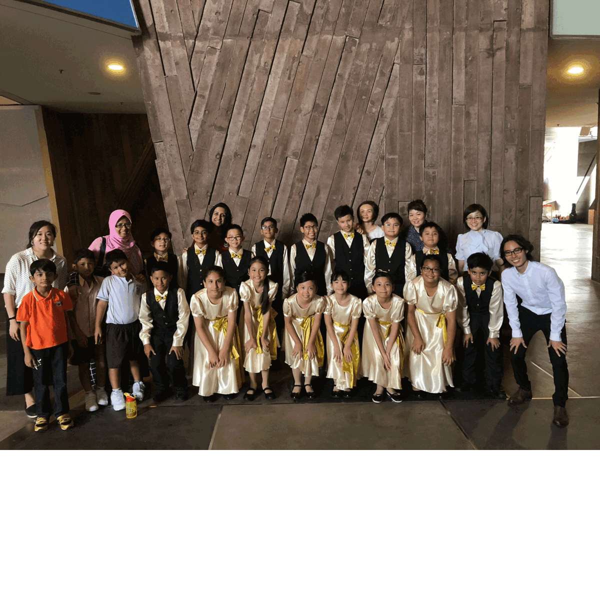 A group of children in matching gold and black performance outfits pose with adults in front of a wooden wall.