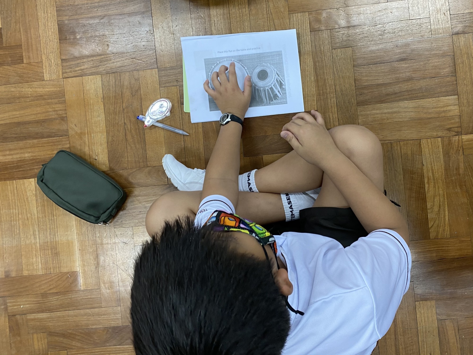 A child in a white shirt sits on a wooden floor, touching a printed sheet showing tabla drums.