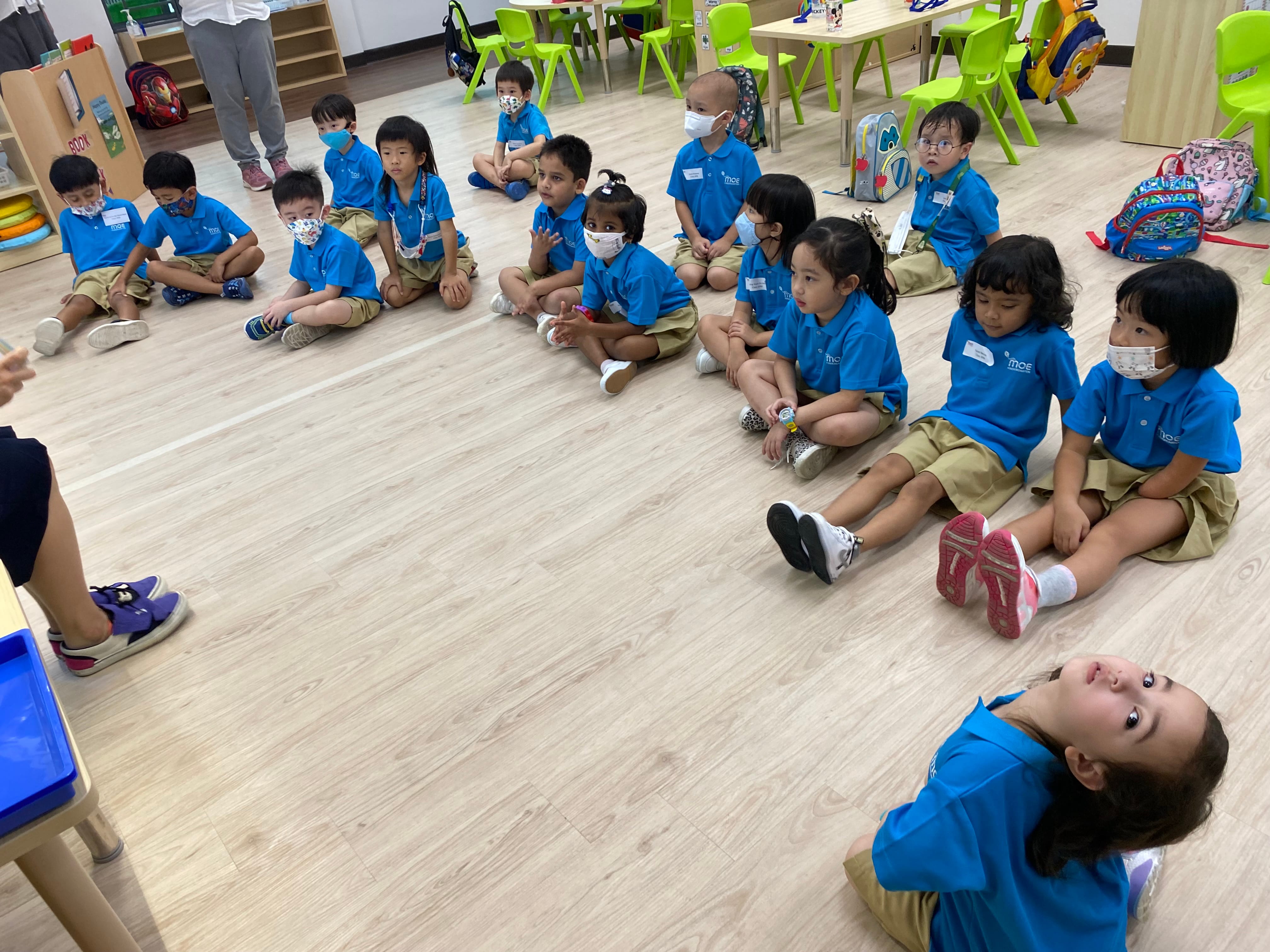 Children in blue uniforms sit on a classroom floor facing a teacher; one child reclines looking up.