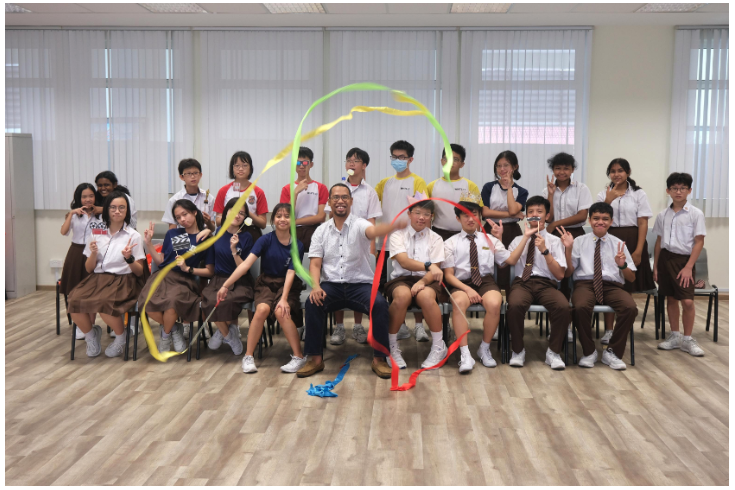 A group of school students and a teacher pose indoors, some waving colorful ribbons in the air.