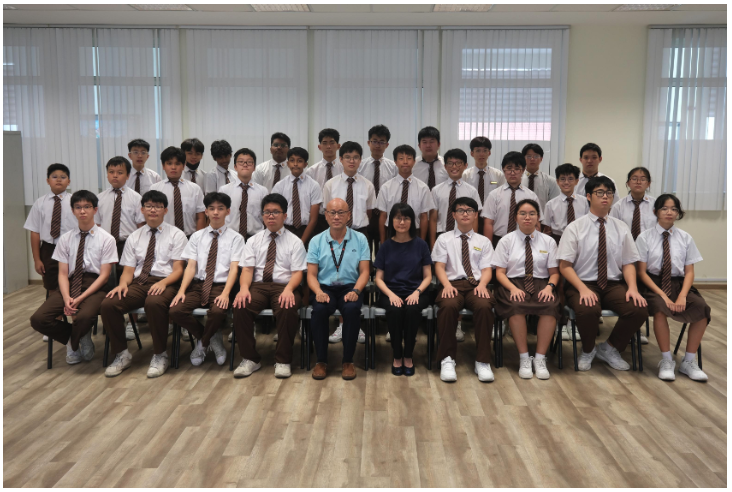 A large group of students in white shirts and brown ties pose with two adults in a classroom setting.