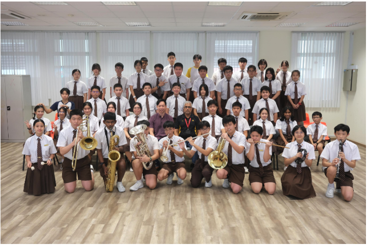 A large group of school students in uniforms pose indoors, some holding brass and woodwind instruments.