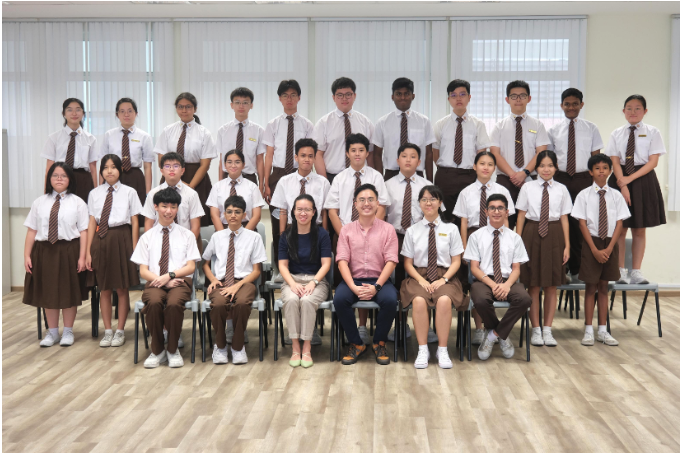 Group photo of students in white shirts and brown ties with two adults seated in the front row indoors.