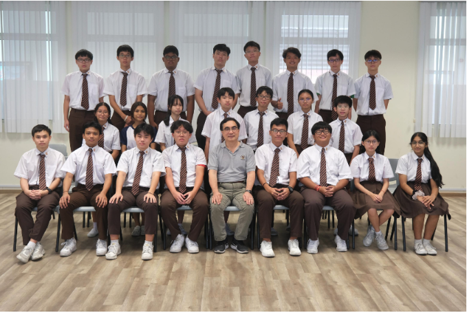 A group of students in white shirts and brown pants with striped ties pose with a teacher in three rows.