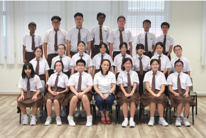 Group of students in white shirts and brown uniforms with striped ties posed in three rows with a teacher seated in front.
