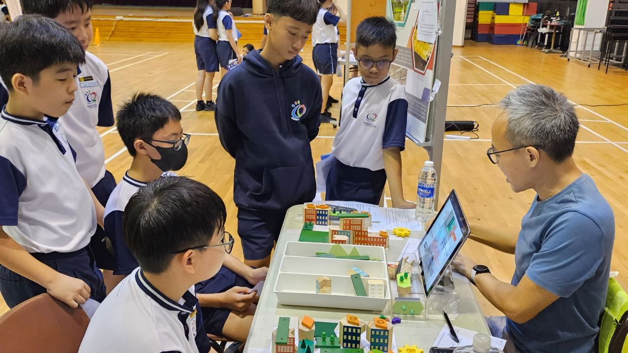 Several school students in uniforms gather around a table with a colorful building model and a laptop in a gymnasium.