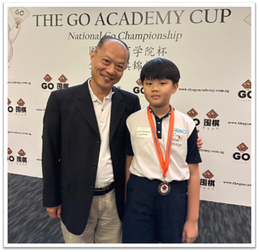An older man and a young boy wearing a medal pose in front of The Go Academy Cup National Go Championship banner.