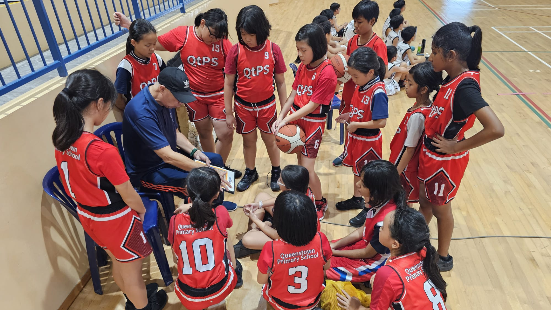A coach seated with a clipboard addresses a group of young girls in red Queenstown Primary School basketball jerseys on a gym floor.