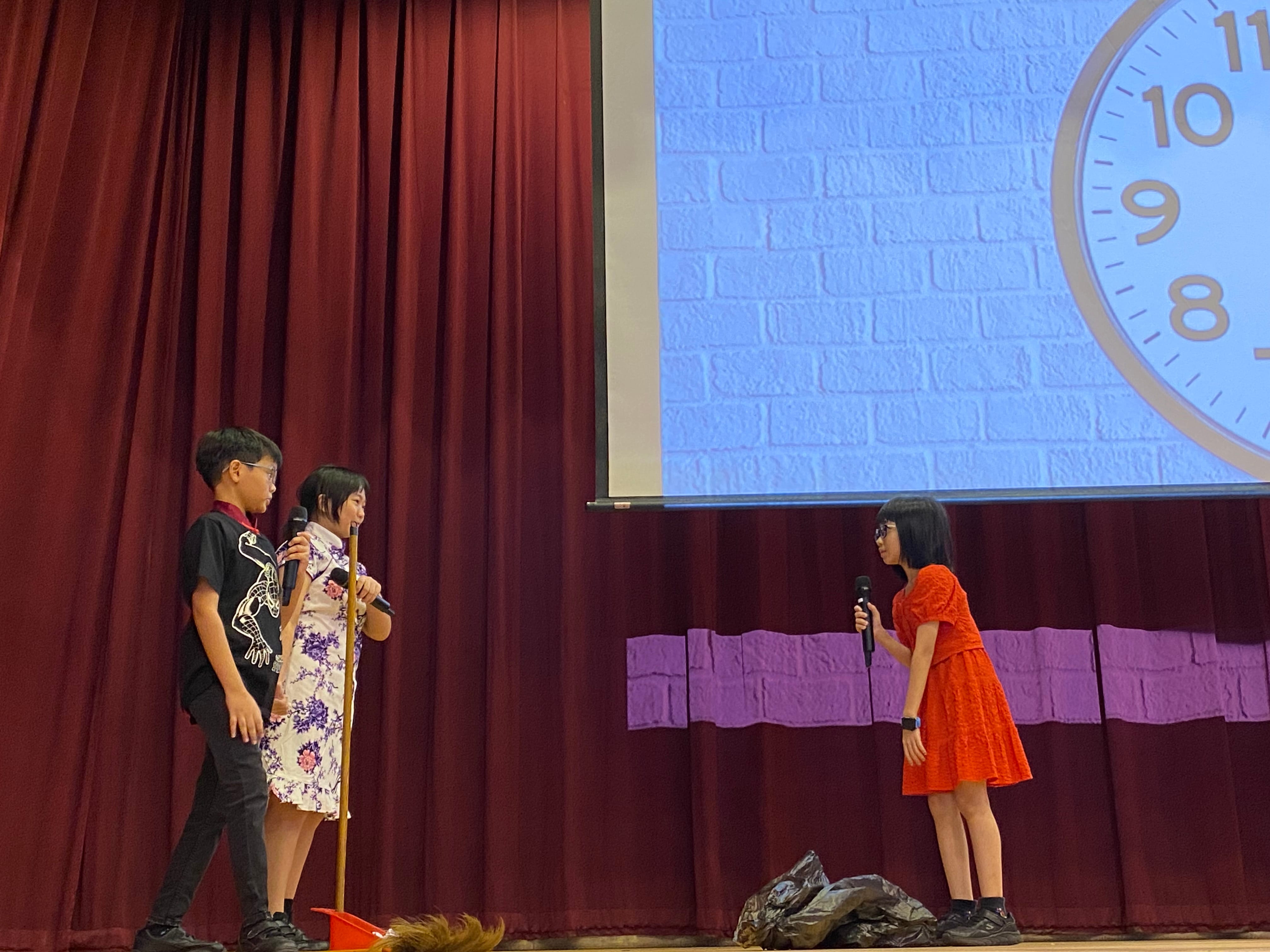 Three children perform on a stage with red curtains, two holding microphones, and a clock projected on a screen behind them.