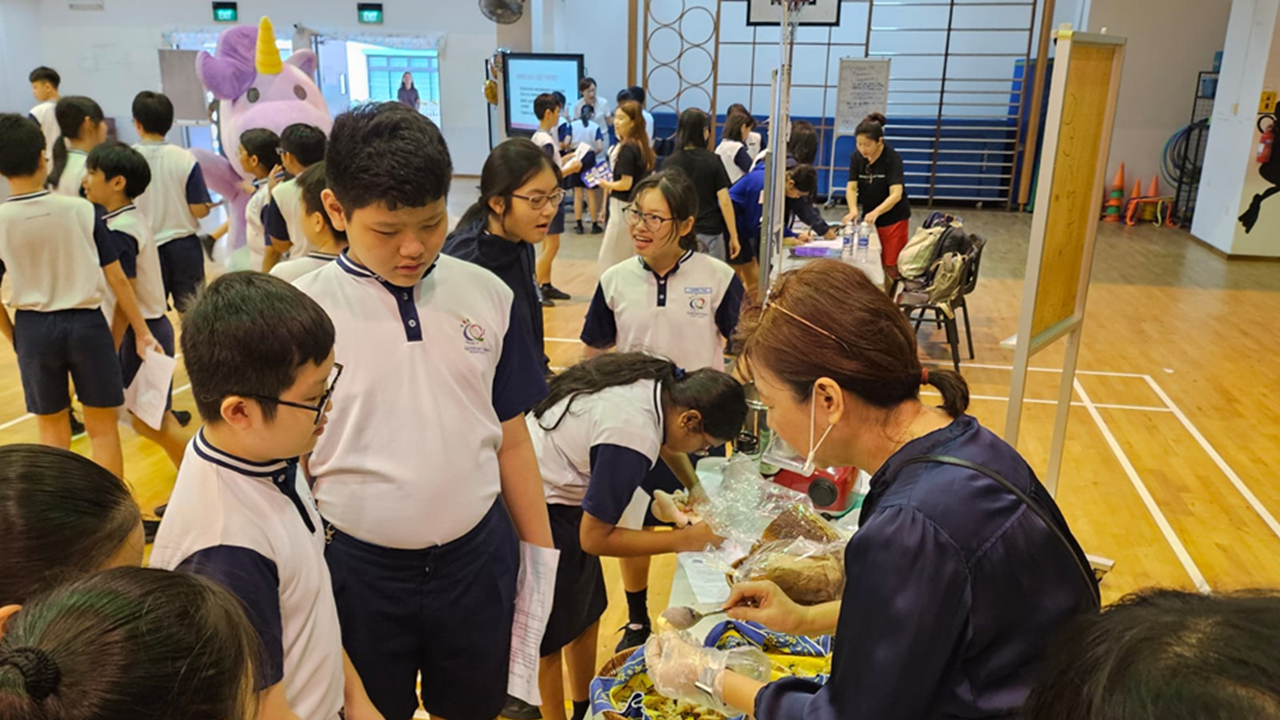 Students in school uniforms gather around a table where an adult serves food in a gym, with a unicorn mascot in the background.