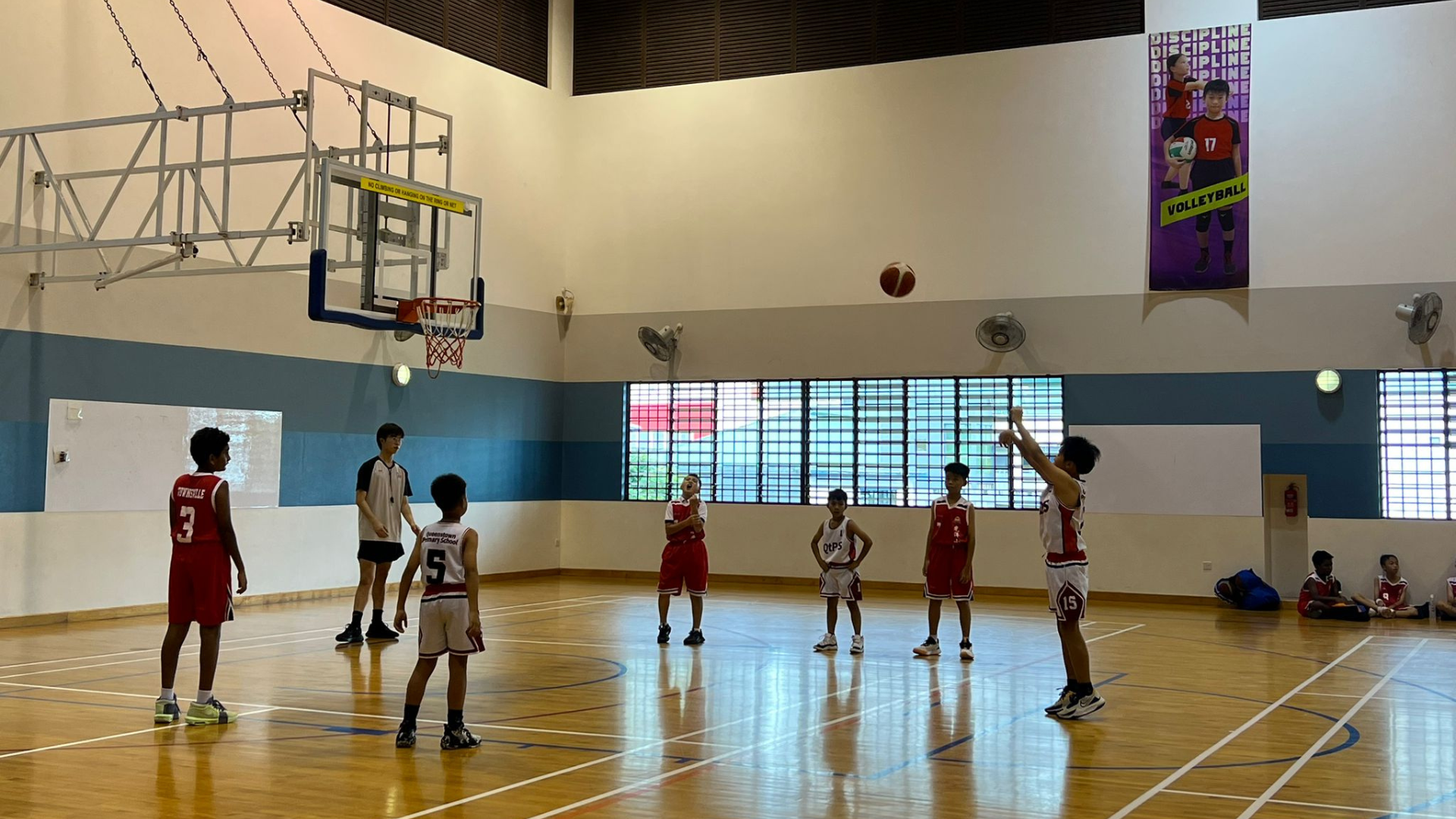 Young boys in red and white uniforms play basketball indoors, with one player shooting toward the hoop.