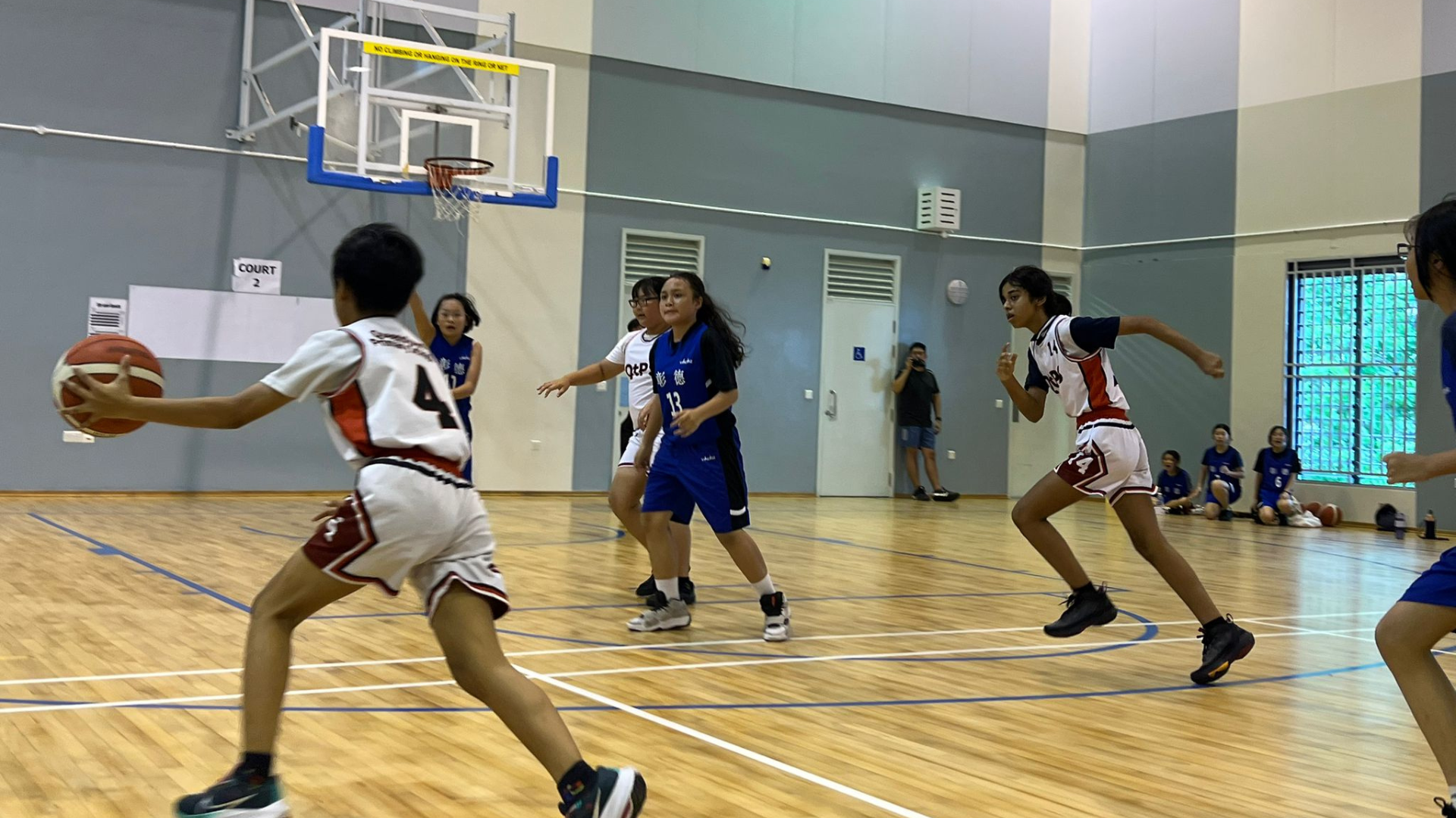 Young players in white and blue uniforms compete in an indoor basketball game on a wooden court.