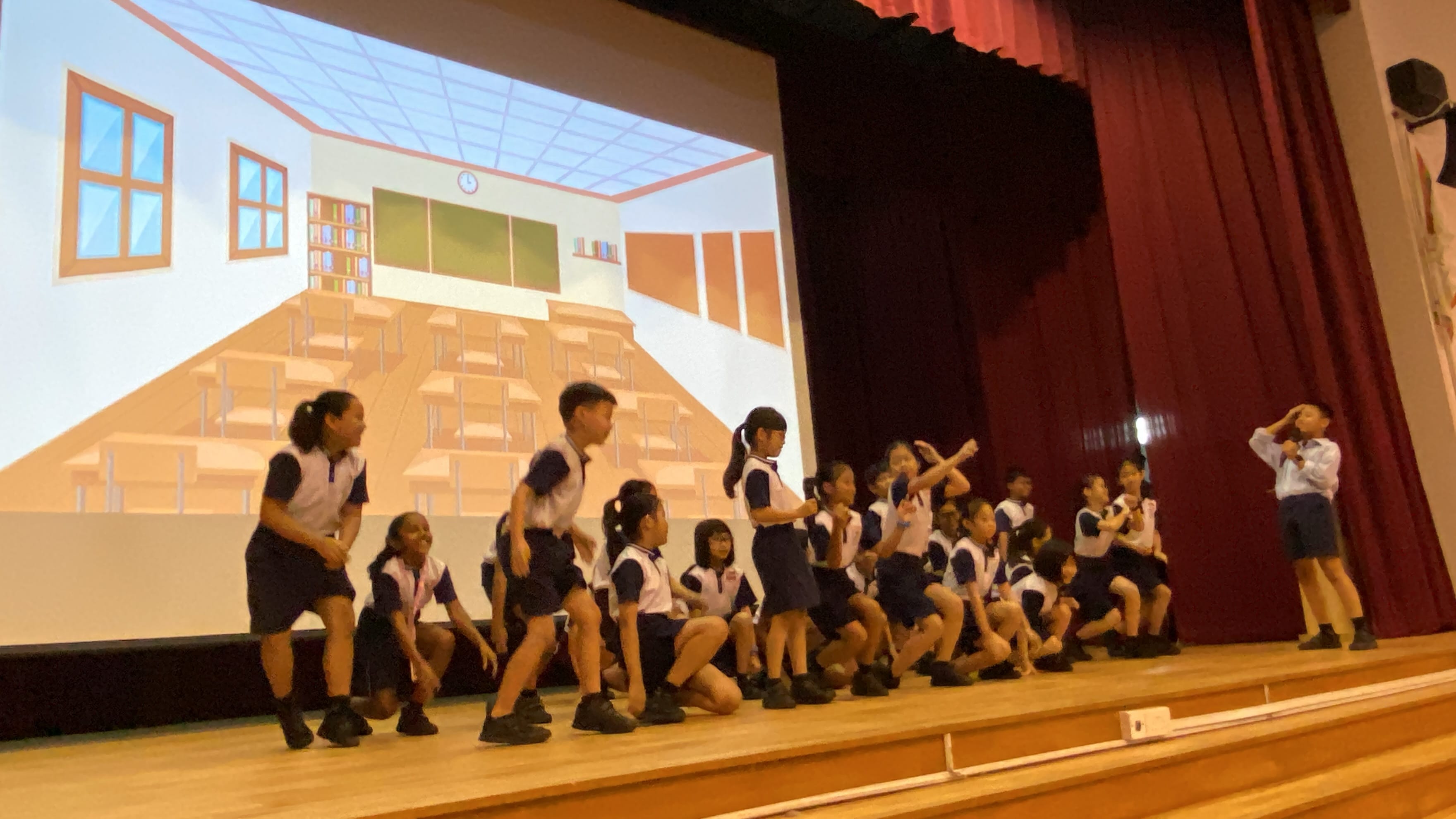 Children in school uniforms perform on a stage in front of a projected cartoon classroom backdrop.