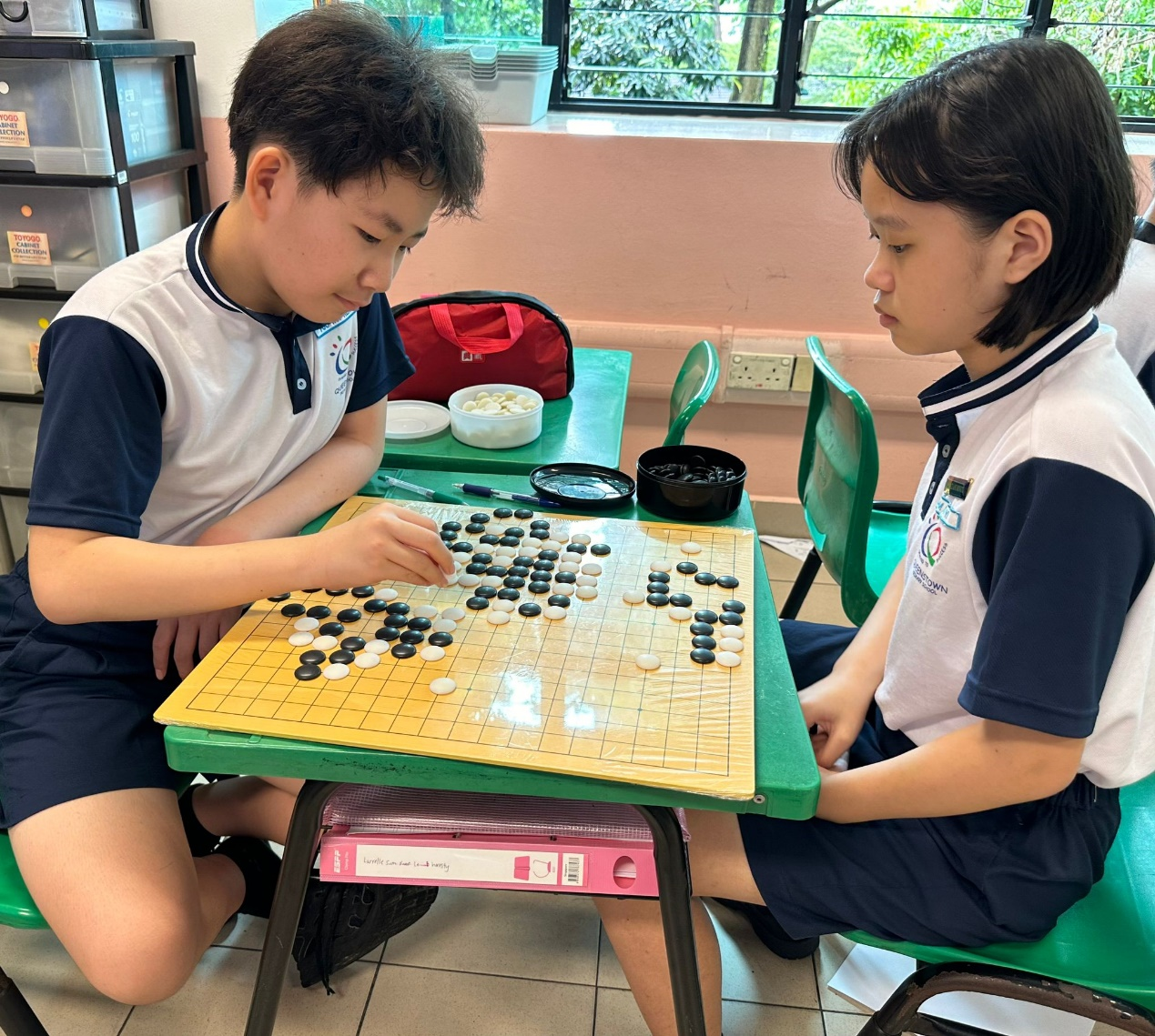 Two children in school uniforms play a game of Go with black and white stones on a yellow board.