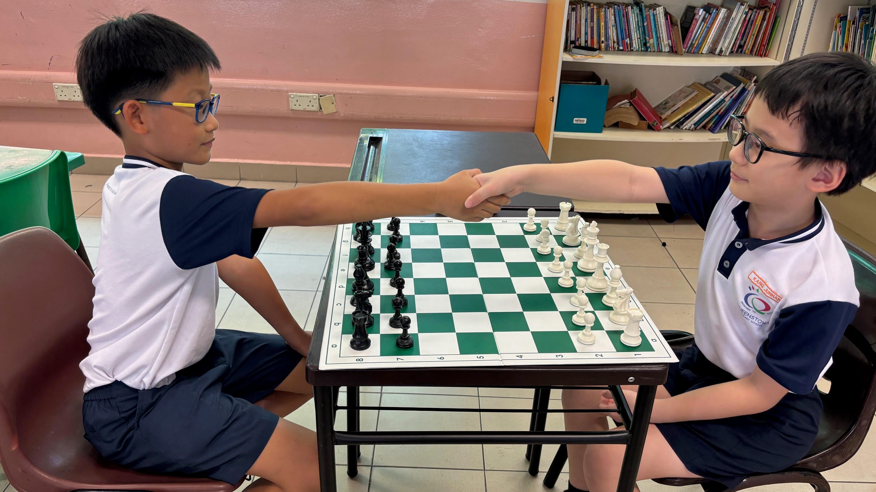 Two boys wearing glasses shake hands over a chess board with pieces mid-game in a classroom.