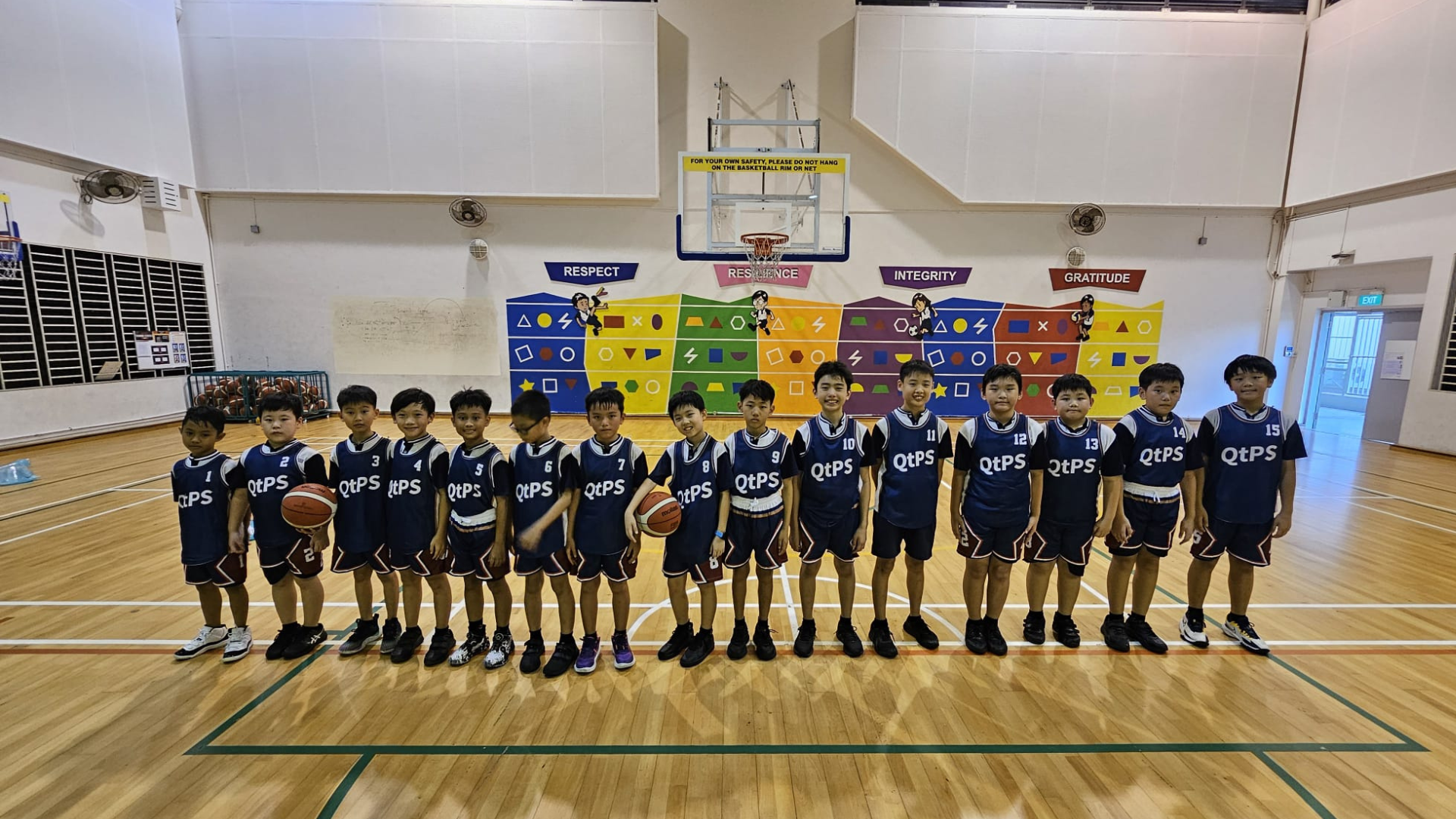Fifteen young boys in navy QtPS basketball uniforms stand in a row on an indoor court beneath a hoop.