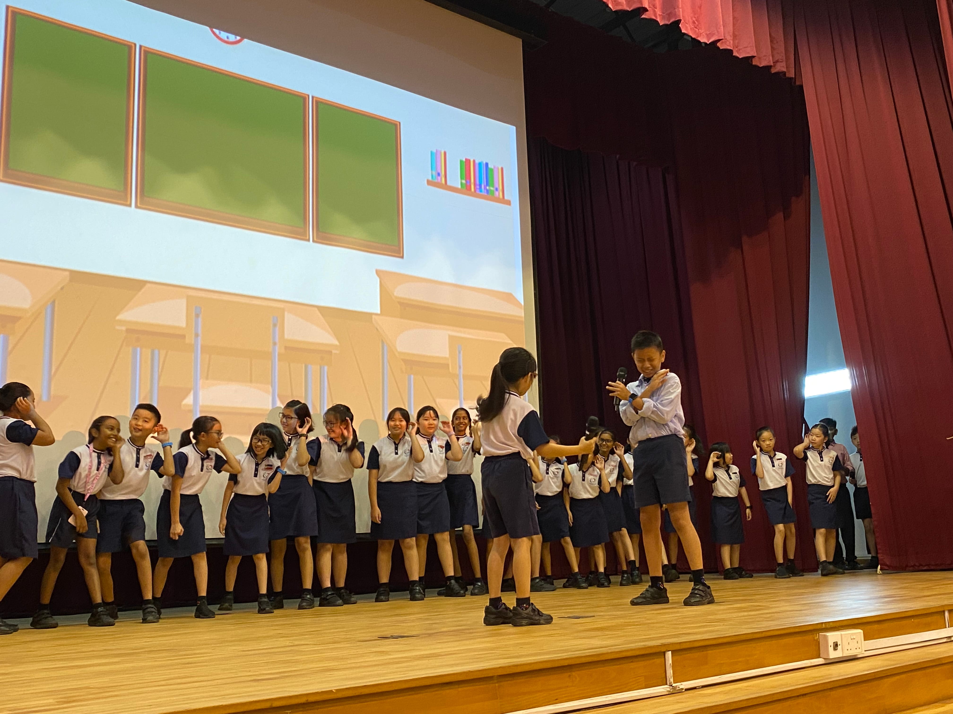 Students in white and navy uniforms perform on a school stage with a cartoon school projected on screen behind them.
