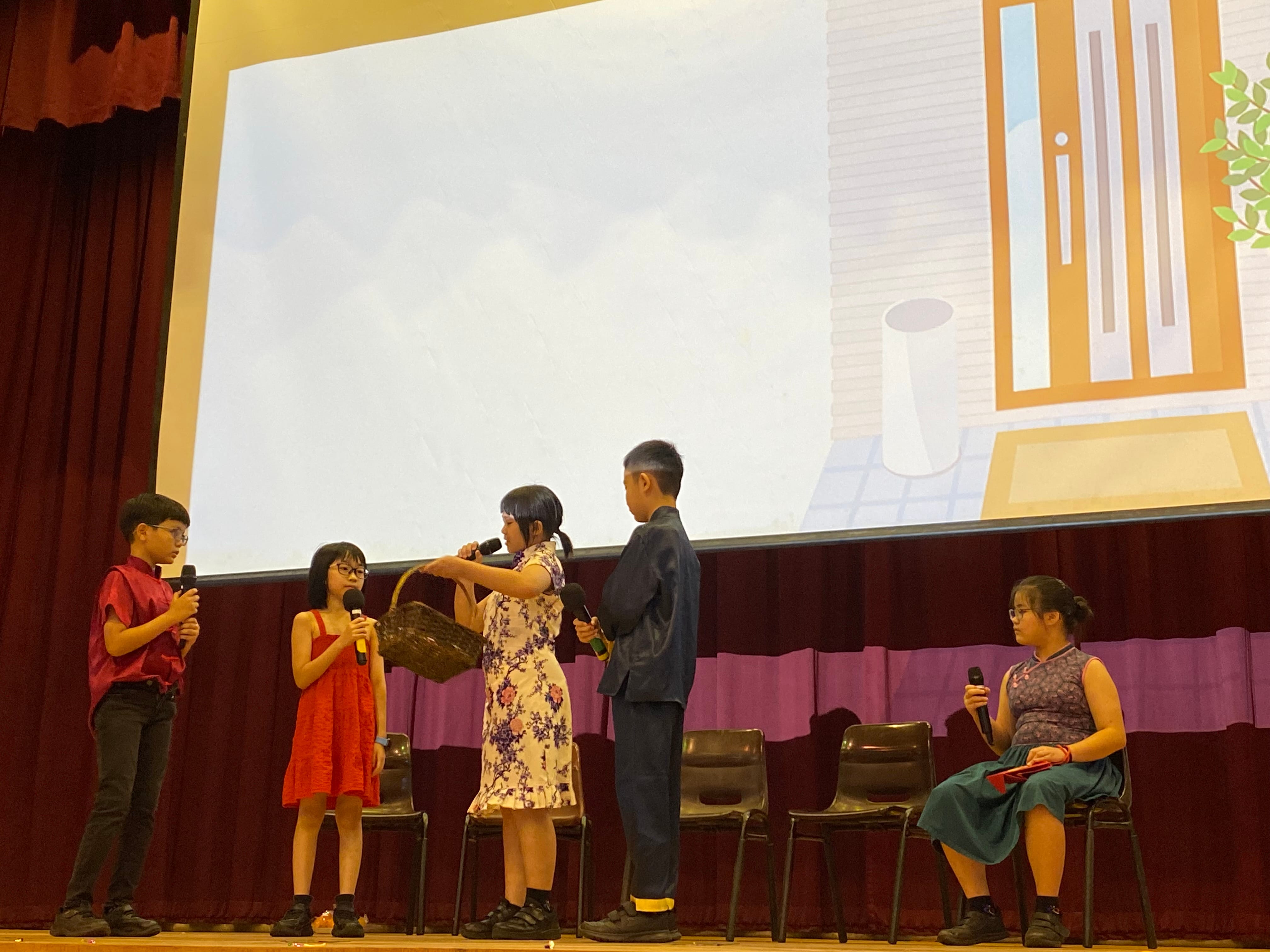 Five children perform on a stage with microphones, one holding a basket, with a projected cartoon backdrop behind them.