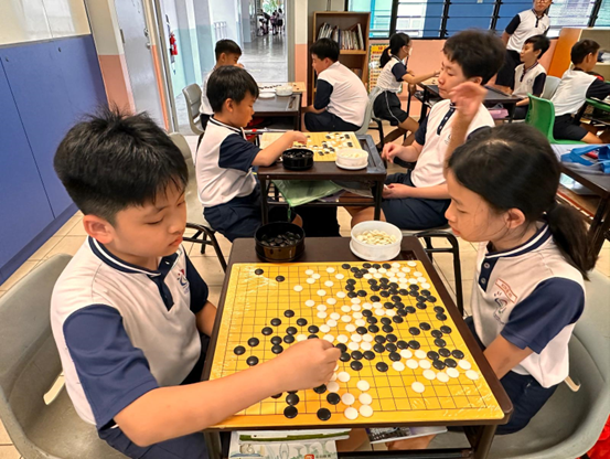 Children in school uniforms playing the board game Go with black and white stones in a classroom.
