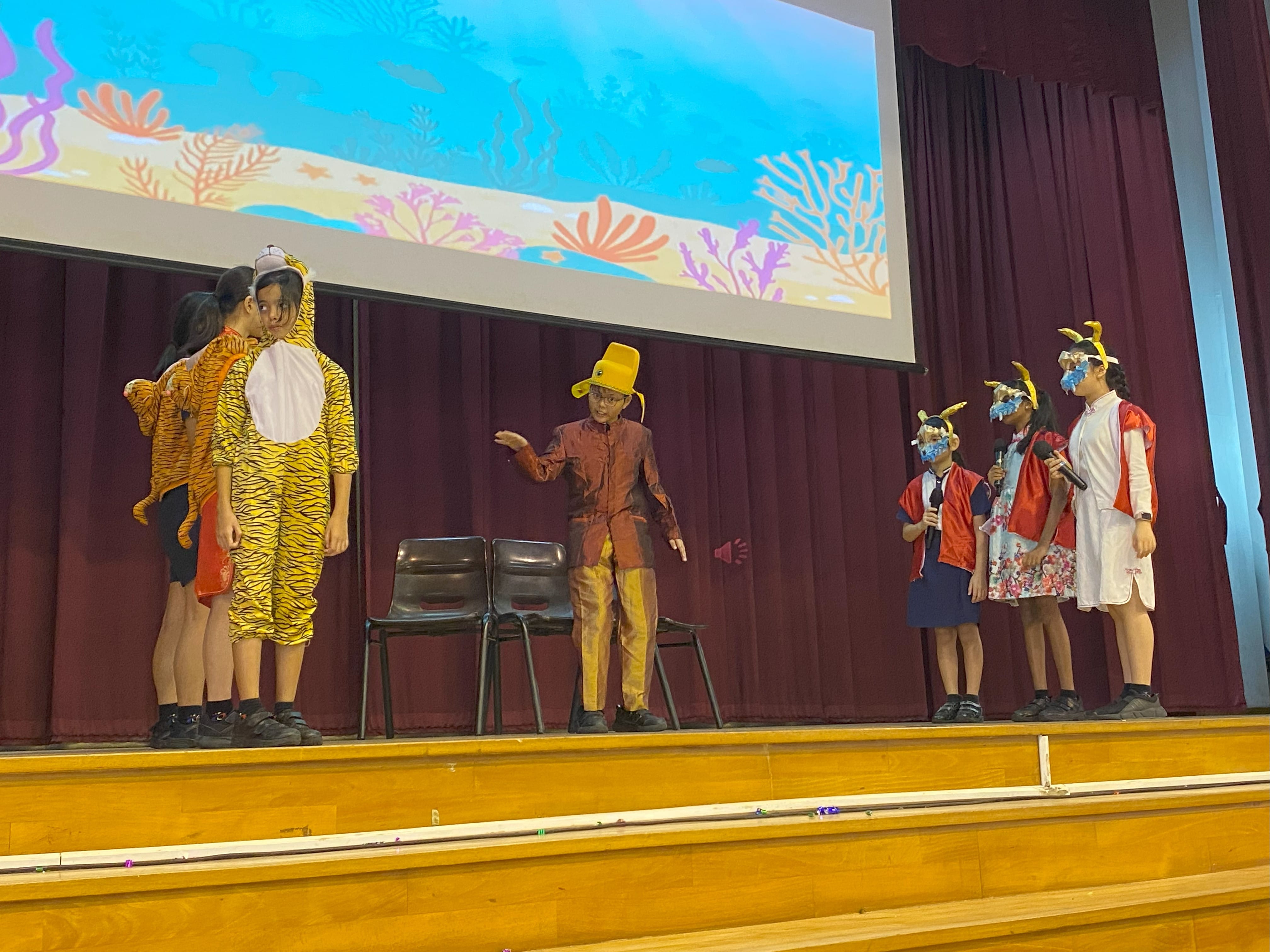 Children in animal costumes perform a school play on stage with an ocean-themed backdrop projected behind them.