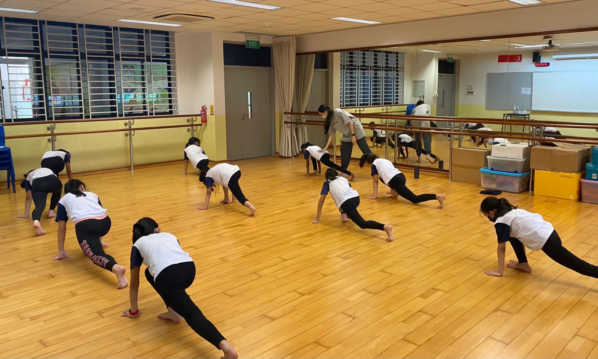 Children in white shirts stretch on a wooden dance studio floor while a teacher guides them near wall mirrors.