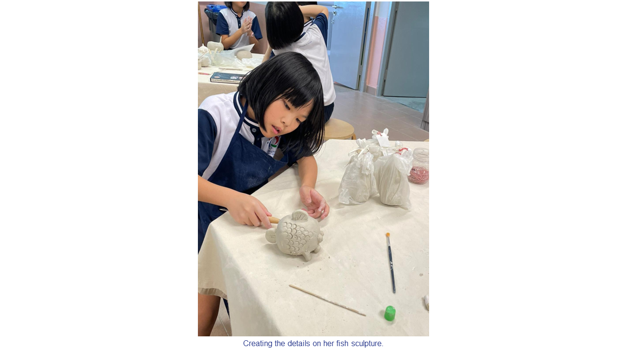 A young girl in a school uniform carefully carves details into a clay fish sculpture on a table.