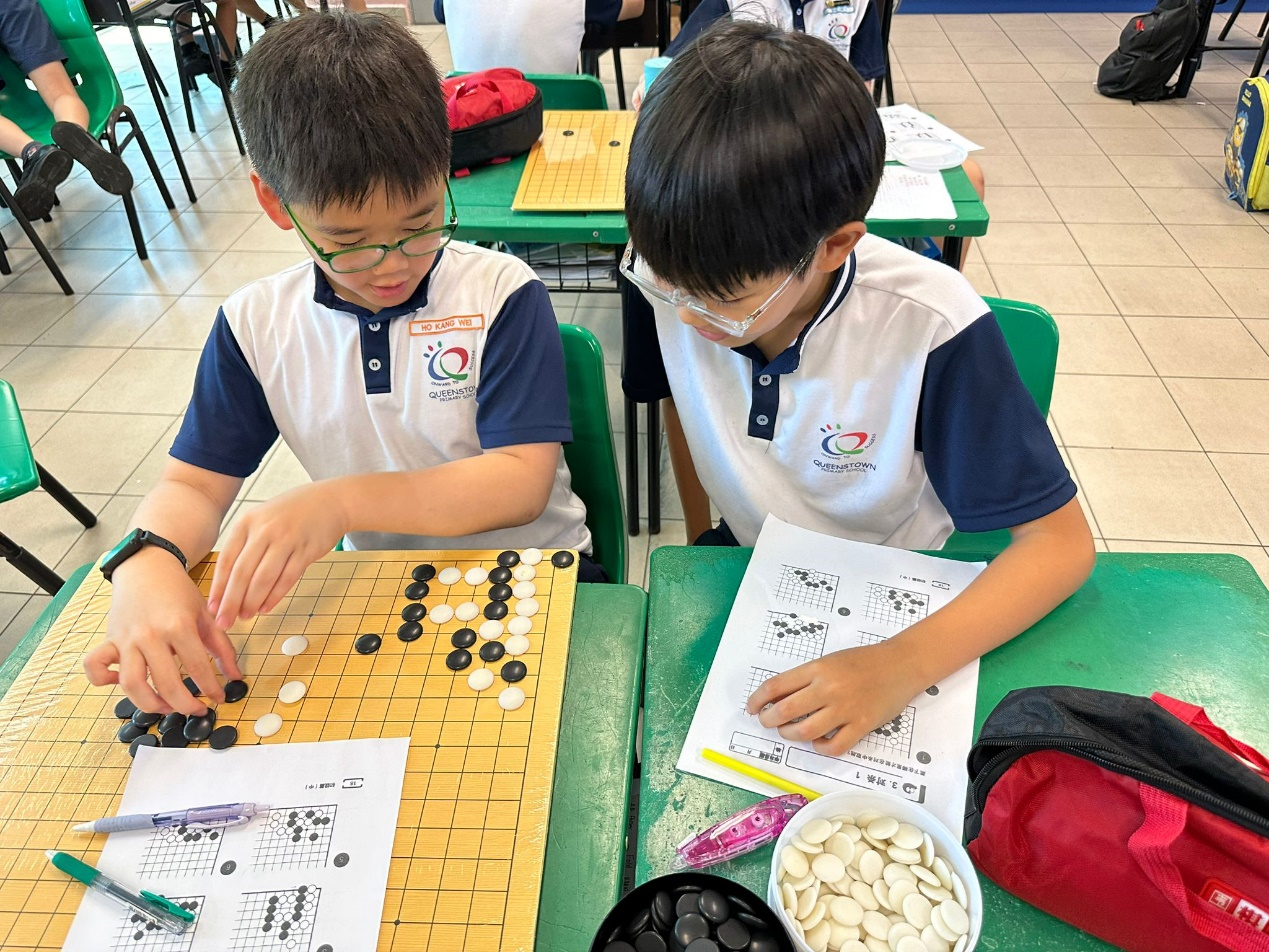 Two young boys in Queenstown school uniforms play a game of Go on a wooden board at green desks.