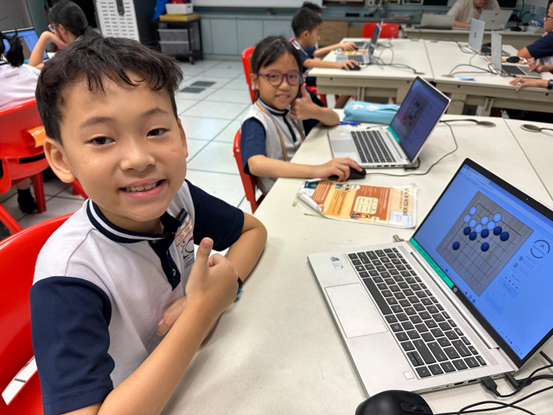 Two young students in uniforms sit at a table with laptops, smiling and giving thumbs up in a classroom.