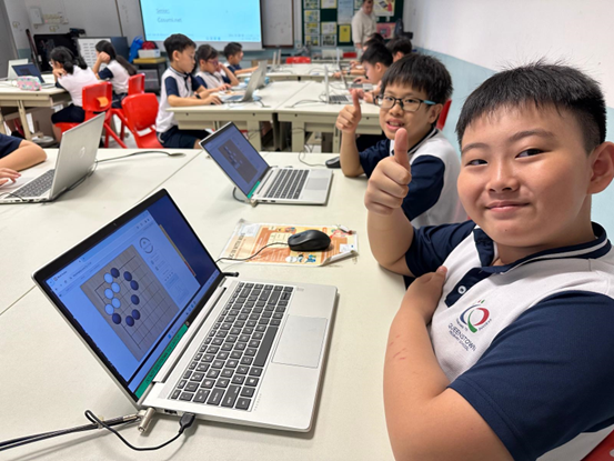 Two smiling students give thumbs up in a classroom while working on laptops showing a grid-based program.
