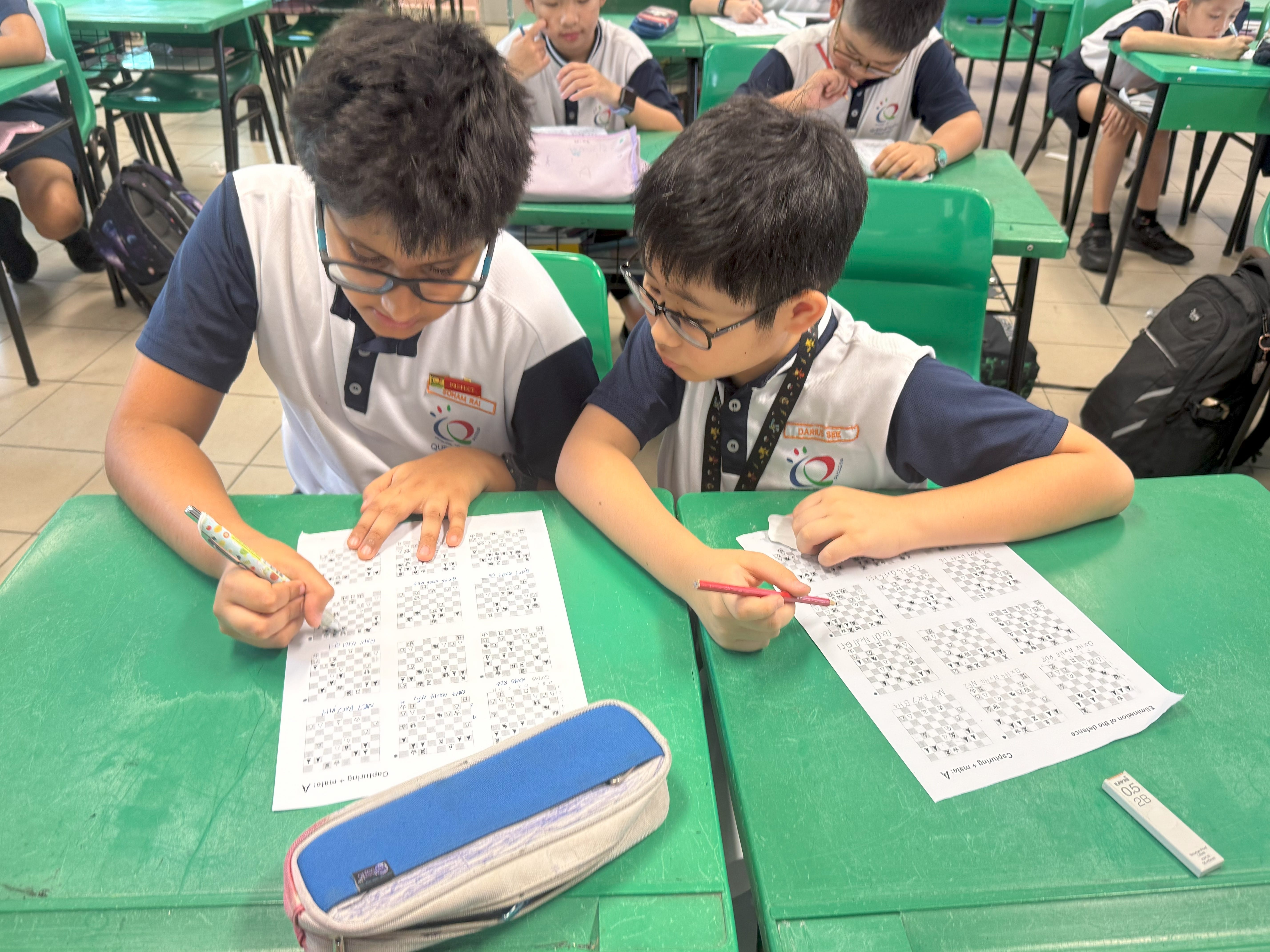 Two boys in school uniforms with glasses work on printed worksheets at green desks in a classroom.