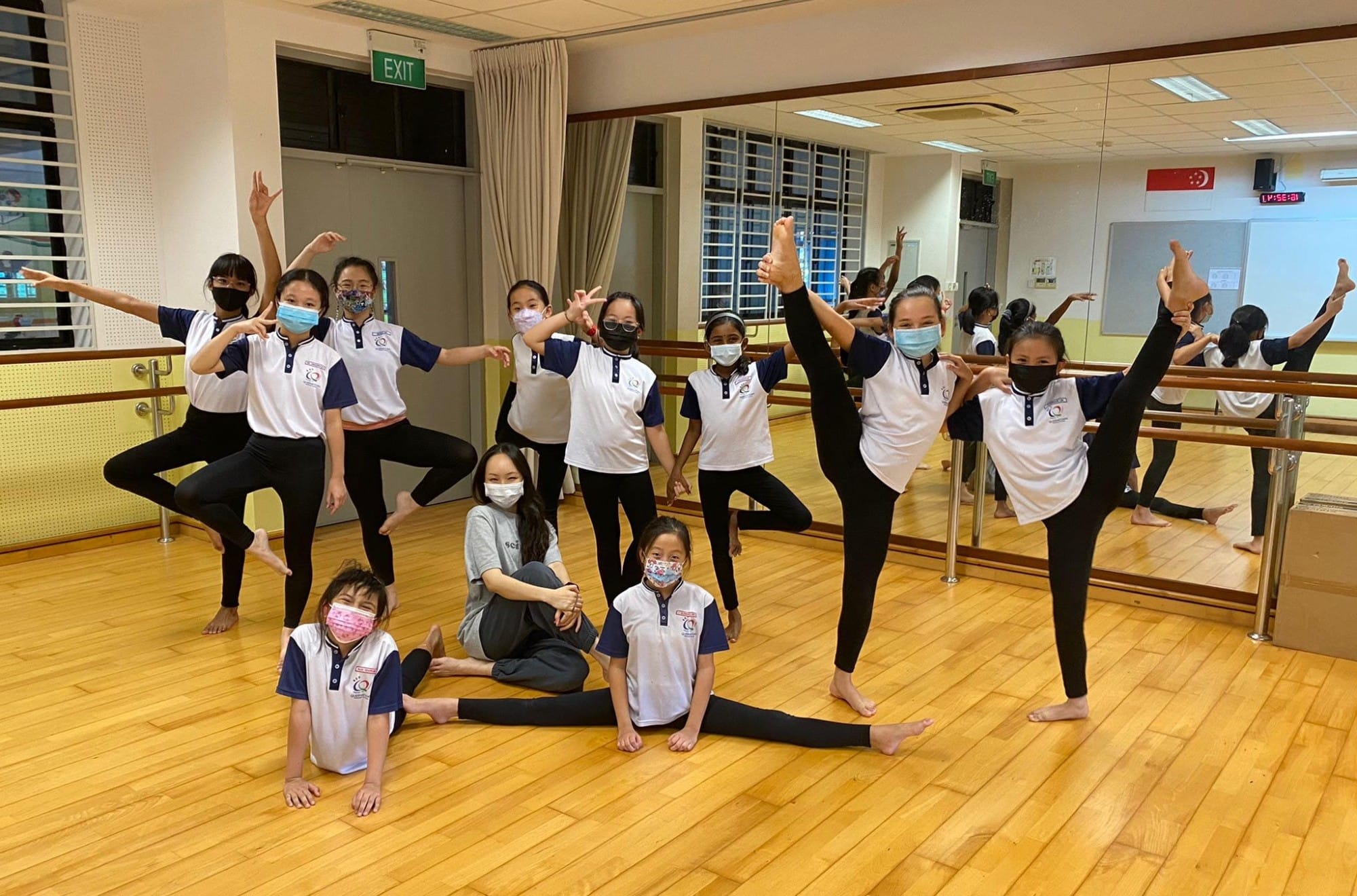 A group of masked schoolgirls in white uniforms pose in various dance and flexibility positions in a studio with mirrors.