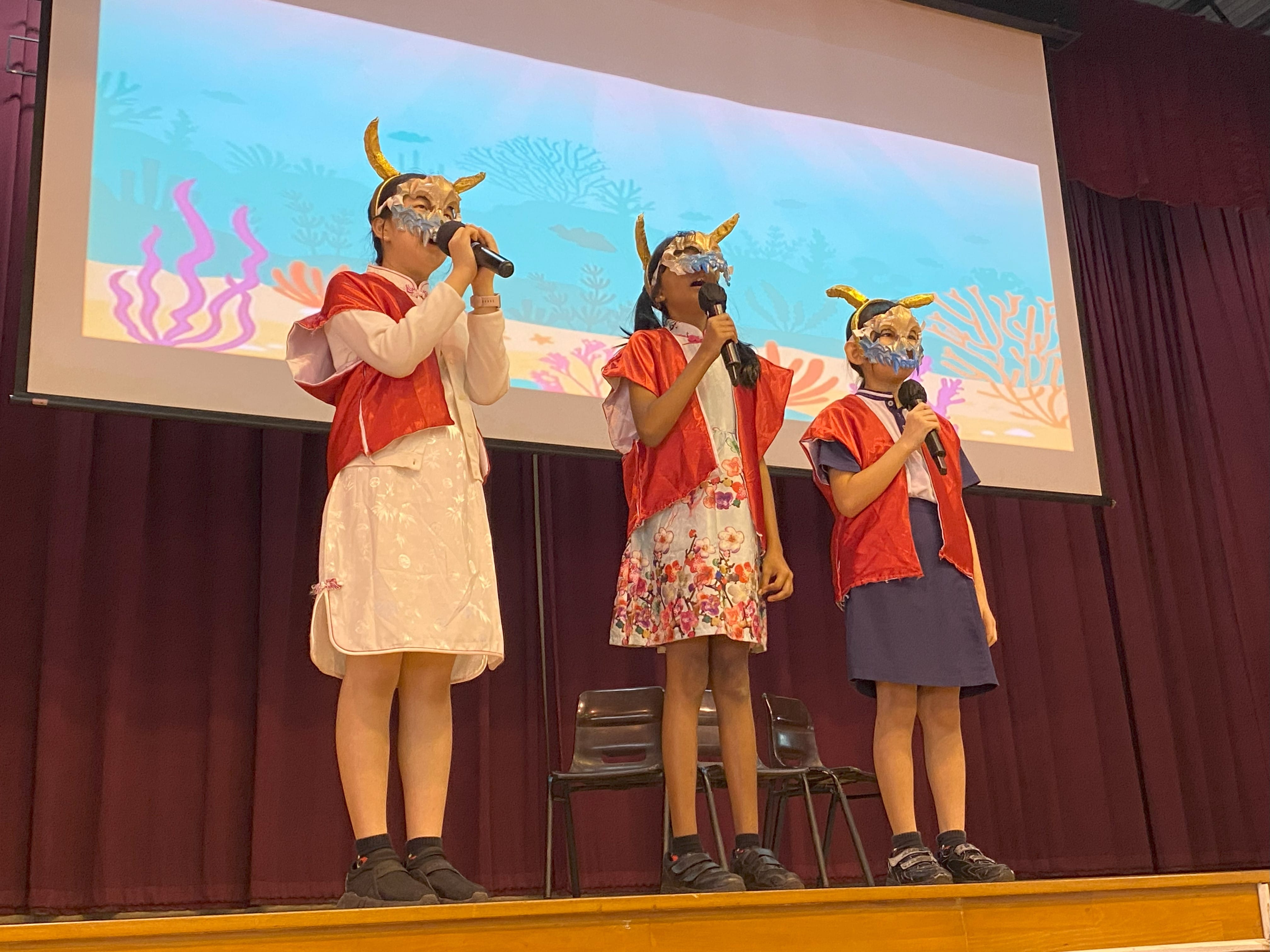 Three children on stage wearing horned masks and red capes, each holding a microphone, with an ocean-themed backdrop.