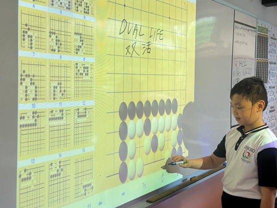 A boy in school uniform points at a projected board showing Go game diagrams labeled "Dual Life" in English and Chinese.