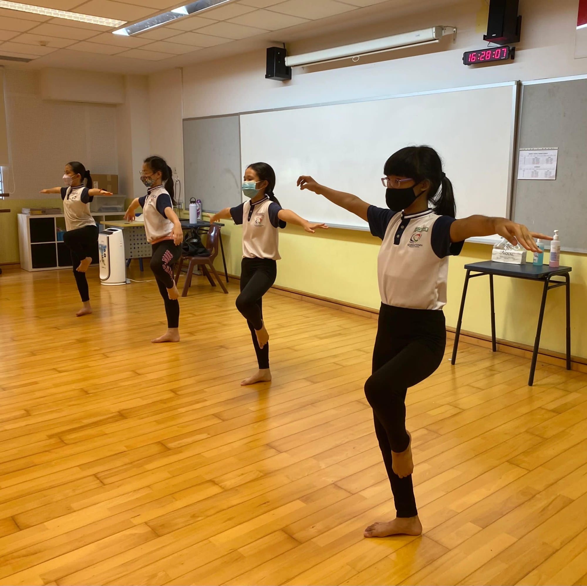 Four masked girls in school uniforms practice a dance pose with arms out and one knee raised on a wooden floor.