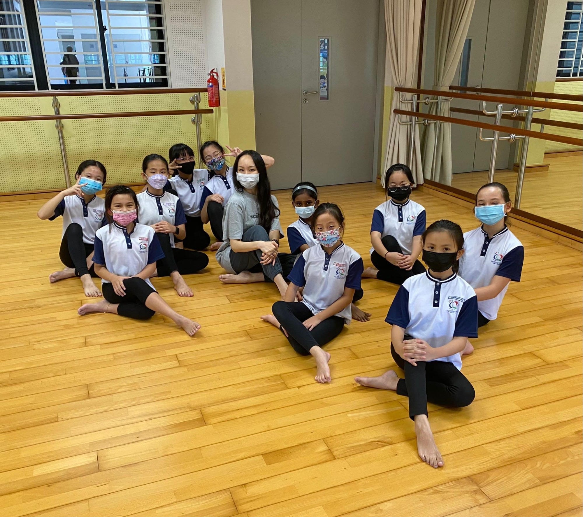 A group of masked children in matching uniforms sit barefoot on a wooden dance studio floor with an adult.