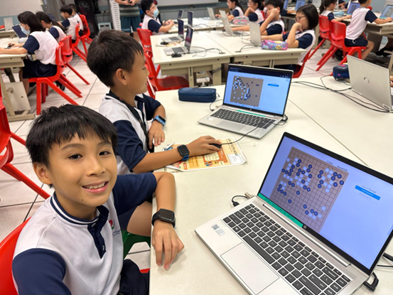 Young students in uniforms sit at tables with laptops displaying a board game, one boy smiling at the camera.