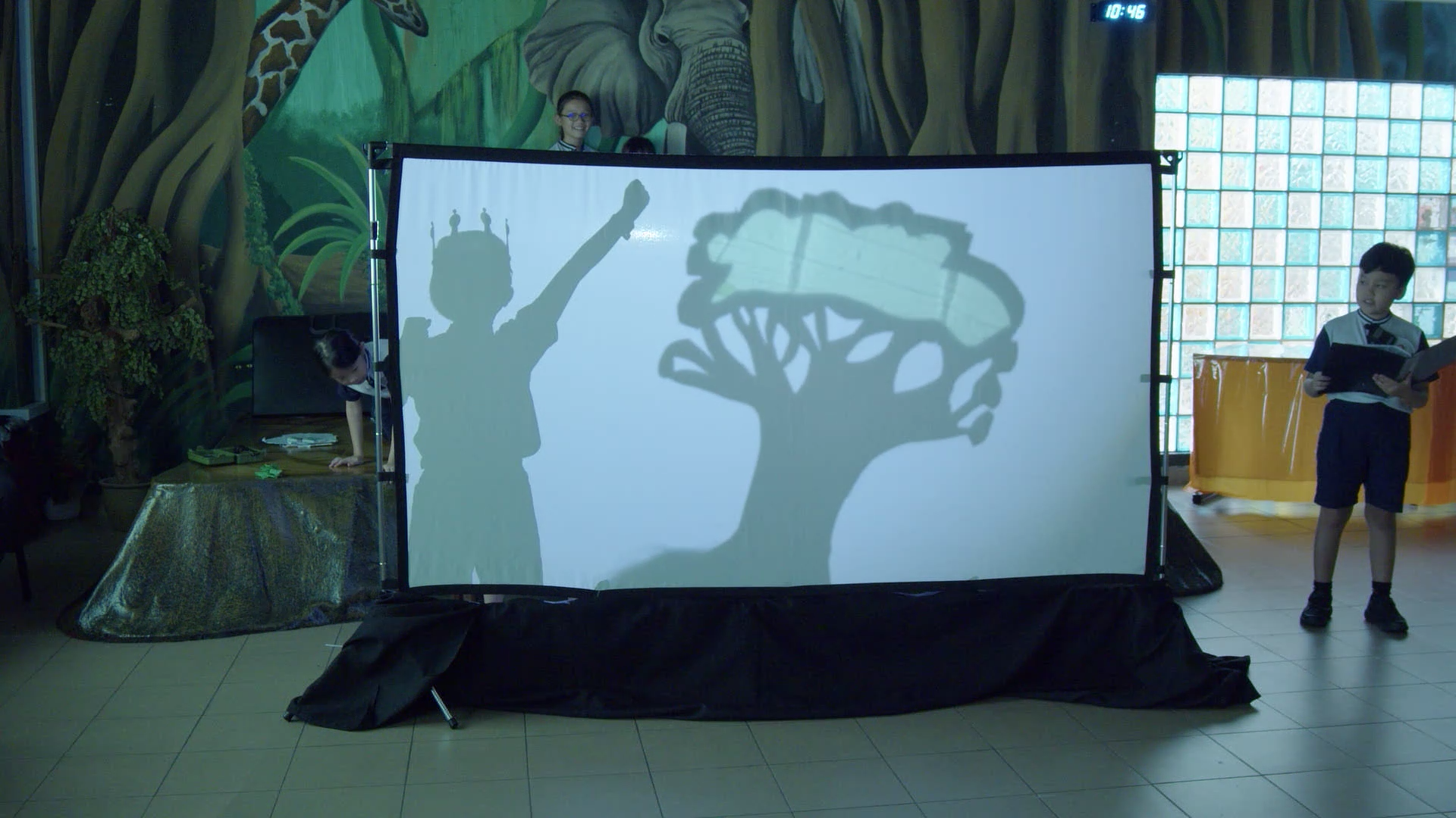 Children perform a shadow puppet show with silhouettes of a crowned figure and tree on a large white screen.