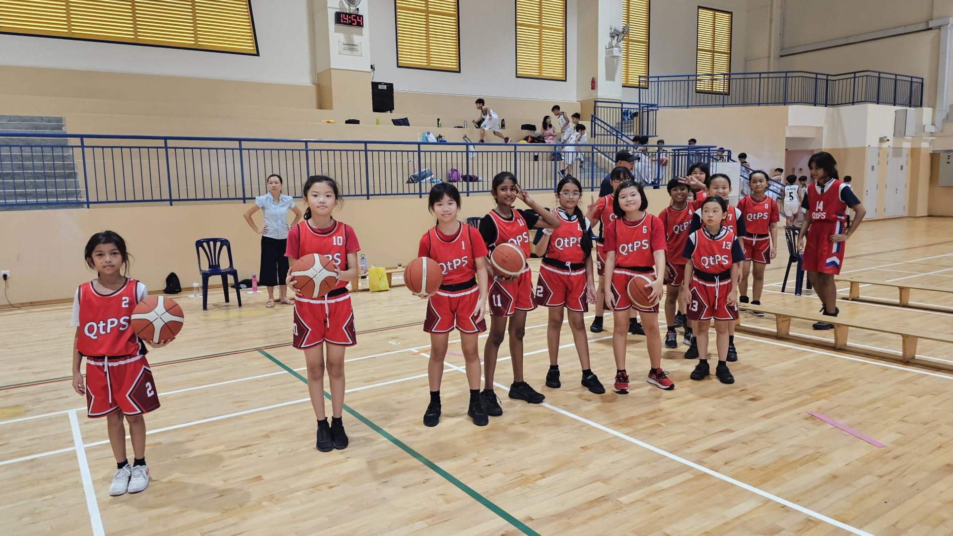 Young girls in red QtPS basketball uniforms stand on an indoor court, several holding basketballs, with a coach behind them.