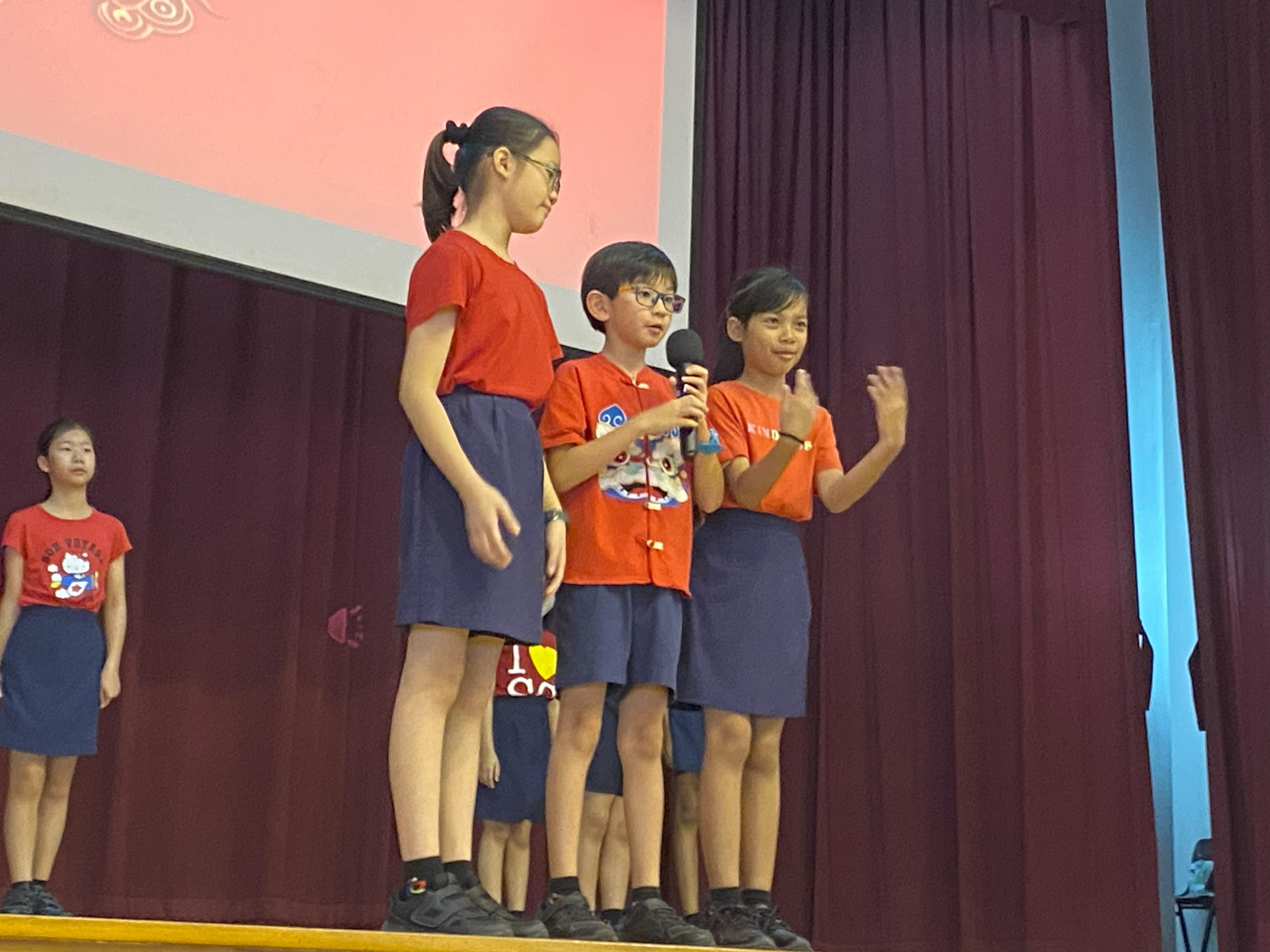 Four children in red shirts and blue shorts stand on a stage, with one boy speaking into a microphone.