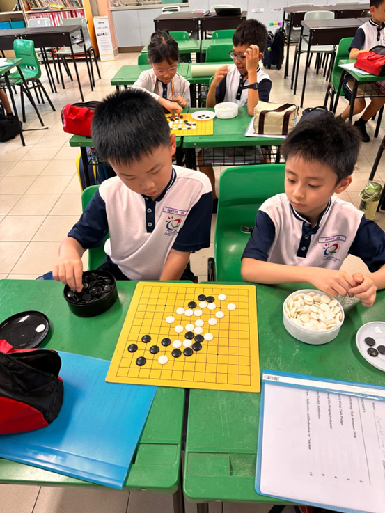 Two young boys in school uniforms play a game of Go on a yellow board at a green desk in a classroom.