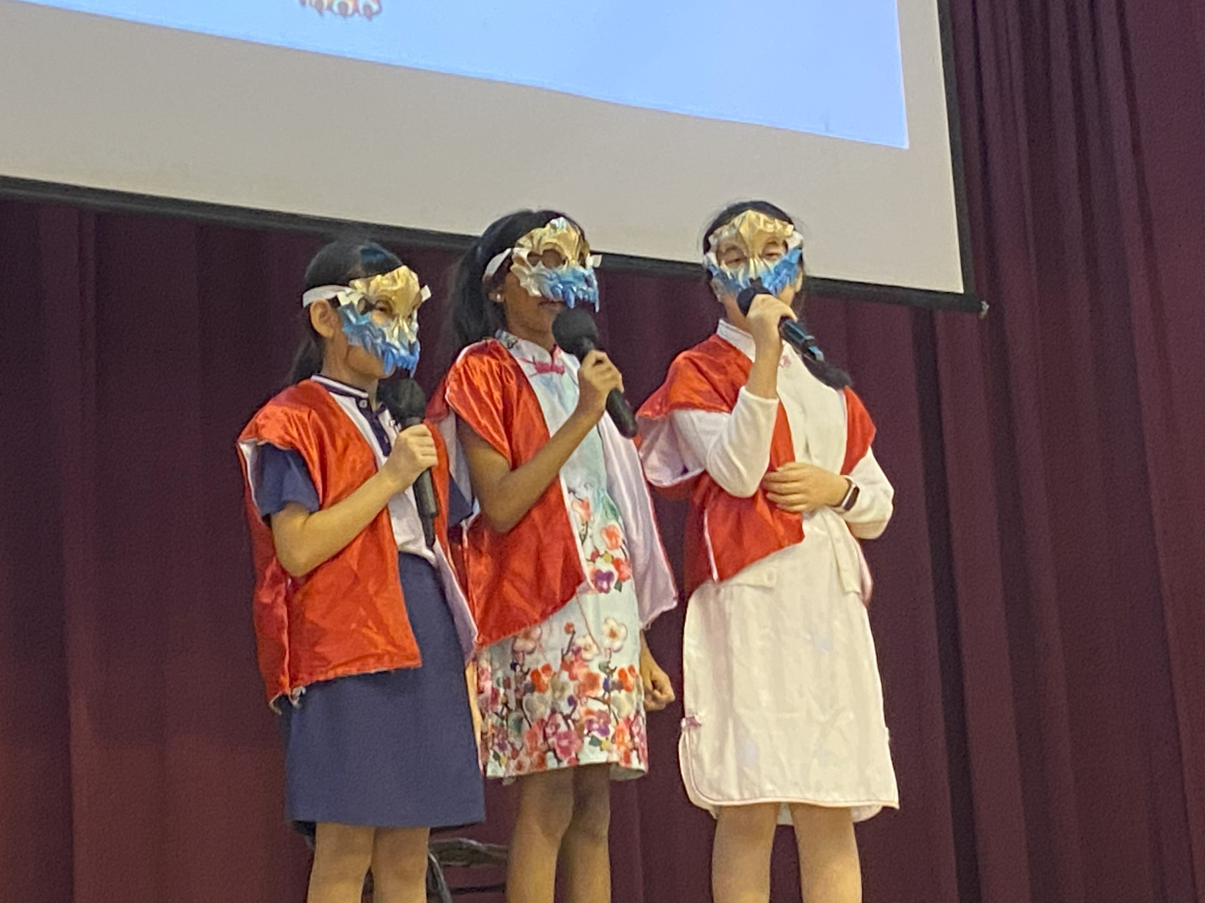 Three girls on stage wearing decorative masks and red capes, each holding a microphone during a school performance.