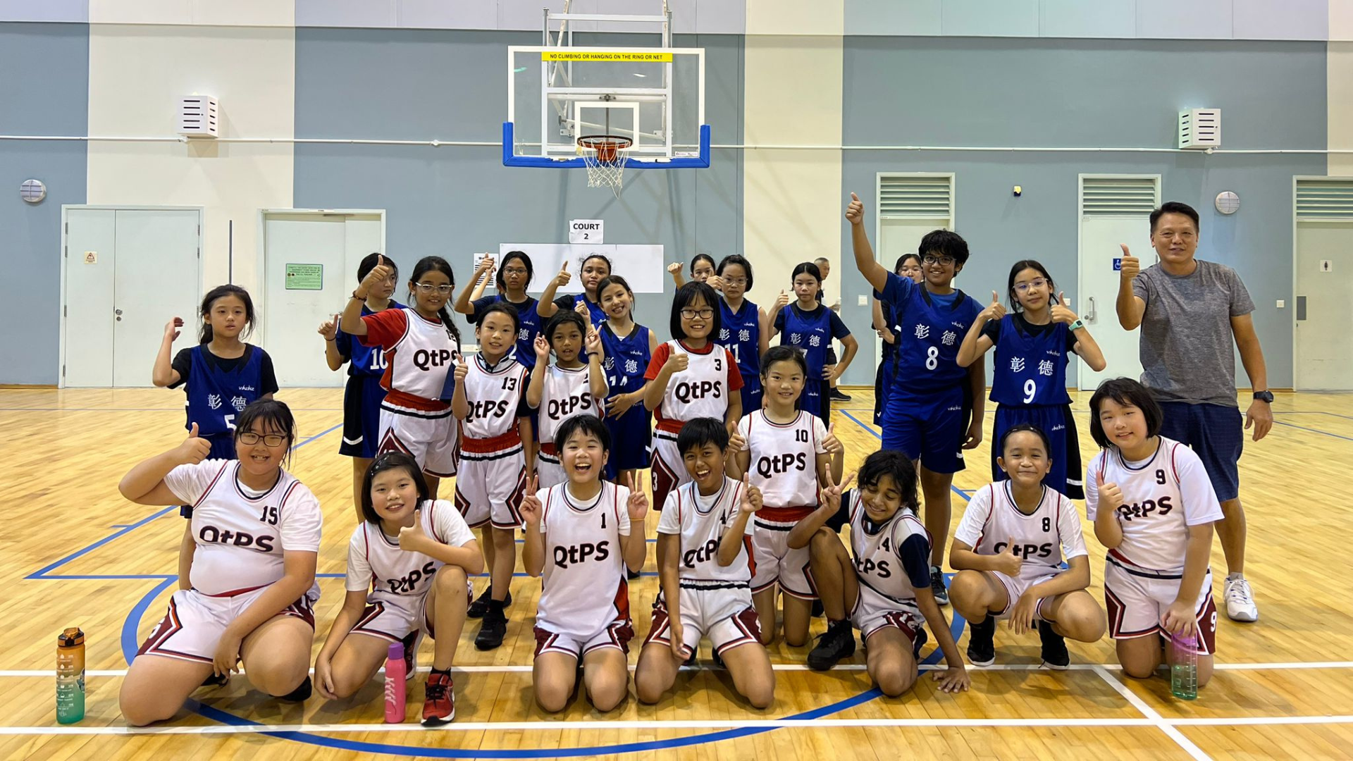 Two youth basketball teams in white QtPS and blue jerseys pose together on an indoor court giving thumbs up.