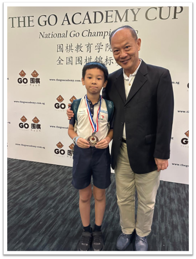 A young boy holding a medal stands with an older man in front of a Go Academy Cup championship banner.