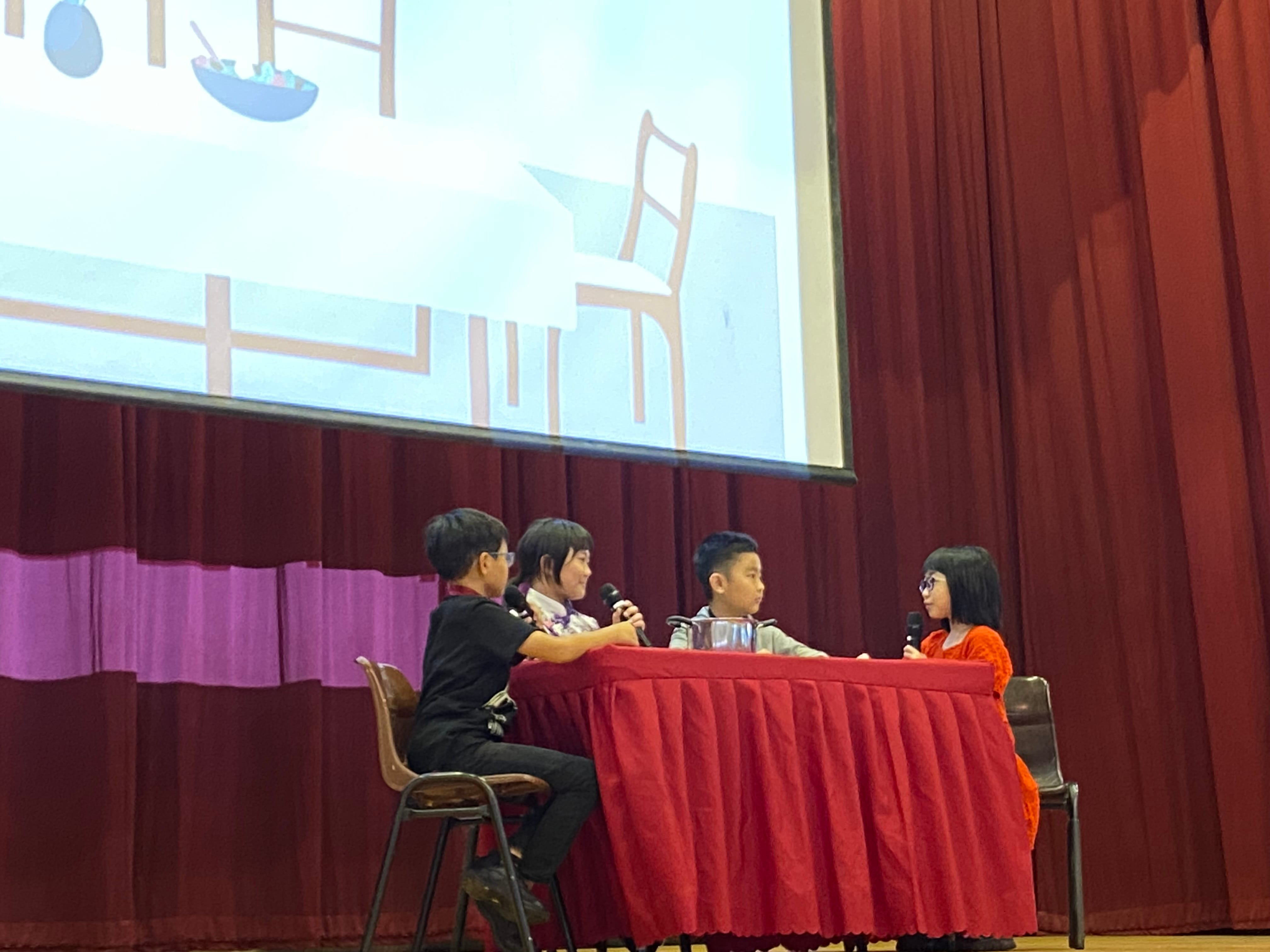 Four children sit at a red-draped table on a stage, holding microphones, with a projected cartoon behind them.