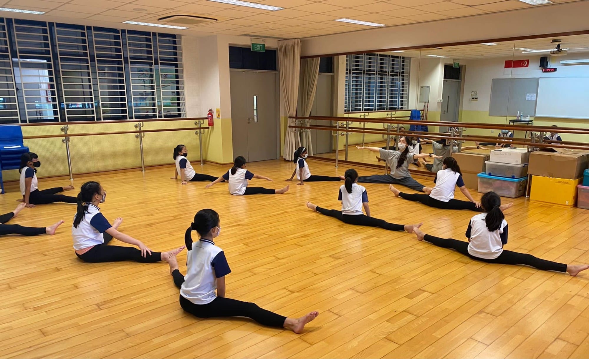 Girls in white shirts and black pants doing splits on a wooden dance studio floor, led by a masked instructor.